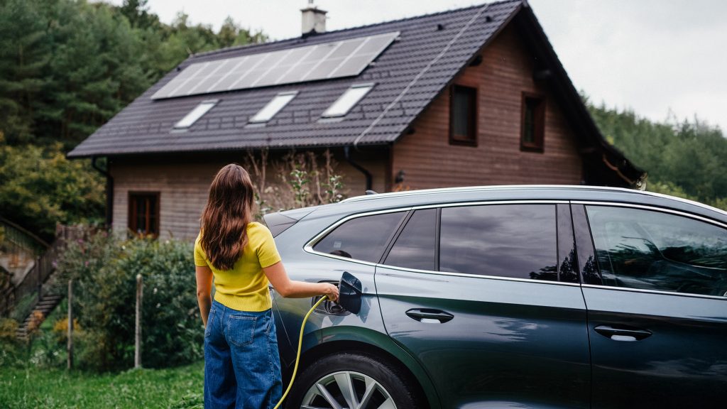 Frau mit Ladekabel am E-Auto vor Haus mit Solarpaneelen auf dem Dach.